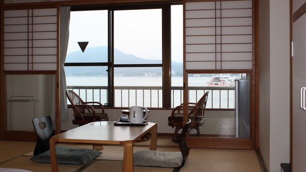 Una habitación japonesa tradicional con suelo de tatami, una mesa baja y sillas. Un gran ventanal ofrece vista a un cuerpo de agua y un barco a lo lejos.