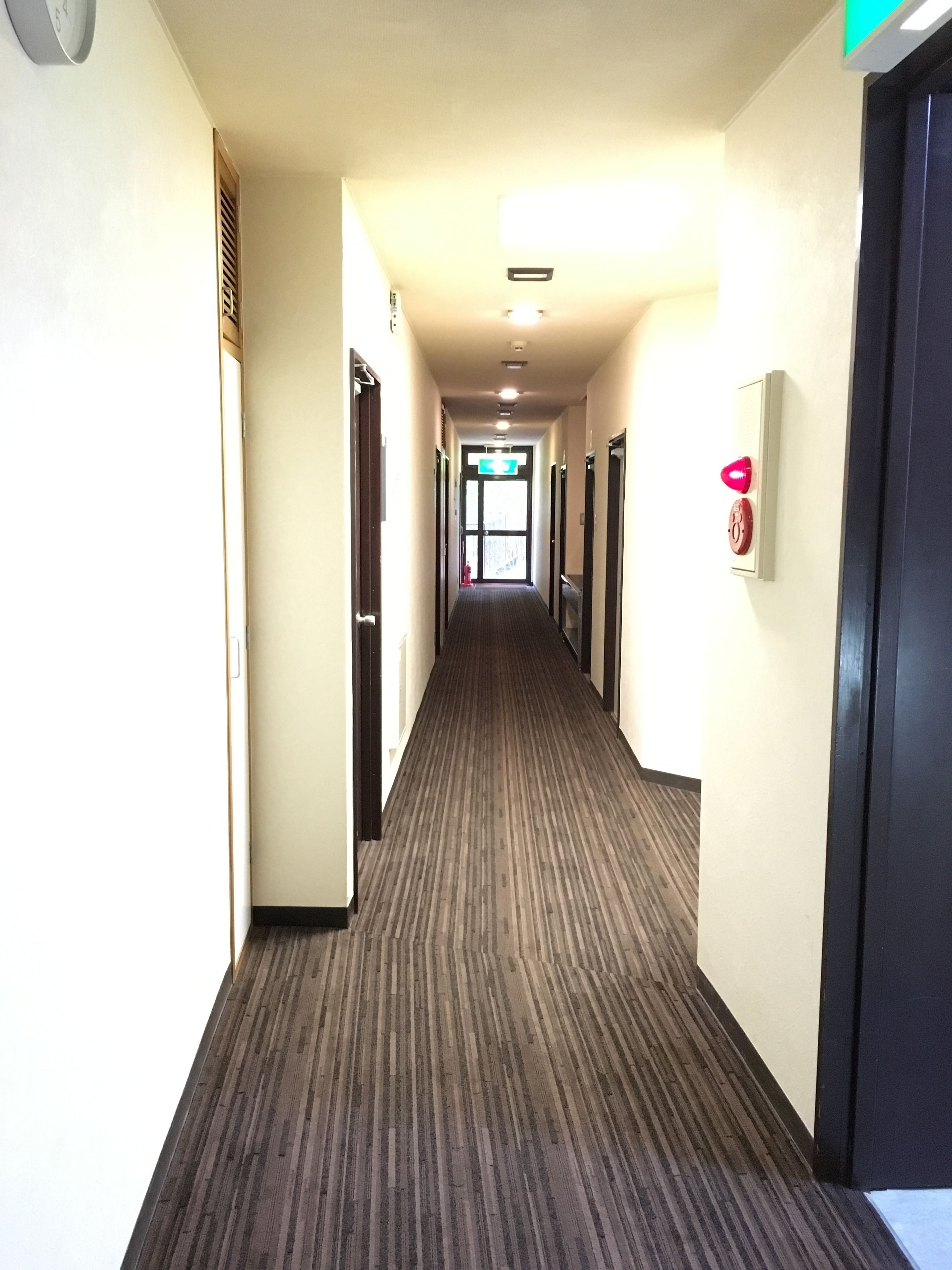 Un couloir d'hôtel avec un sol recouvert de moquette, des portes des deux côtés et un détecteur d'incendie sur le mur de droite.