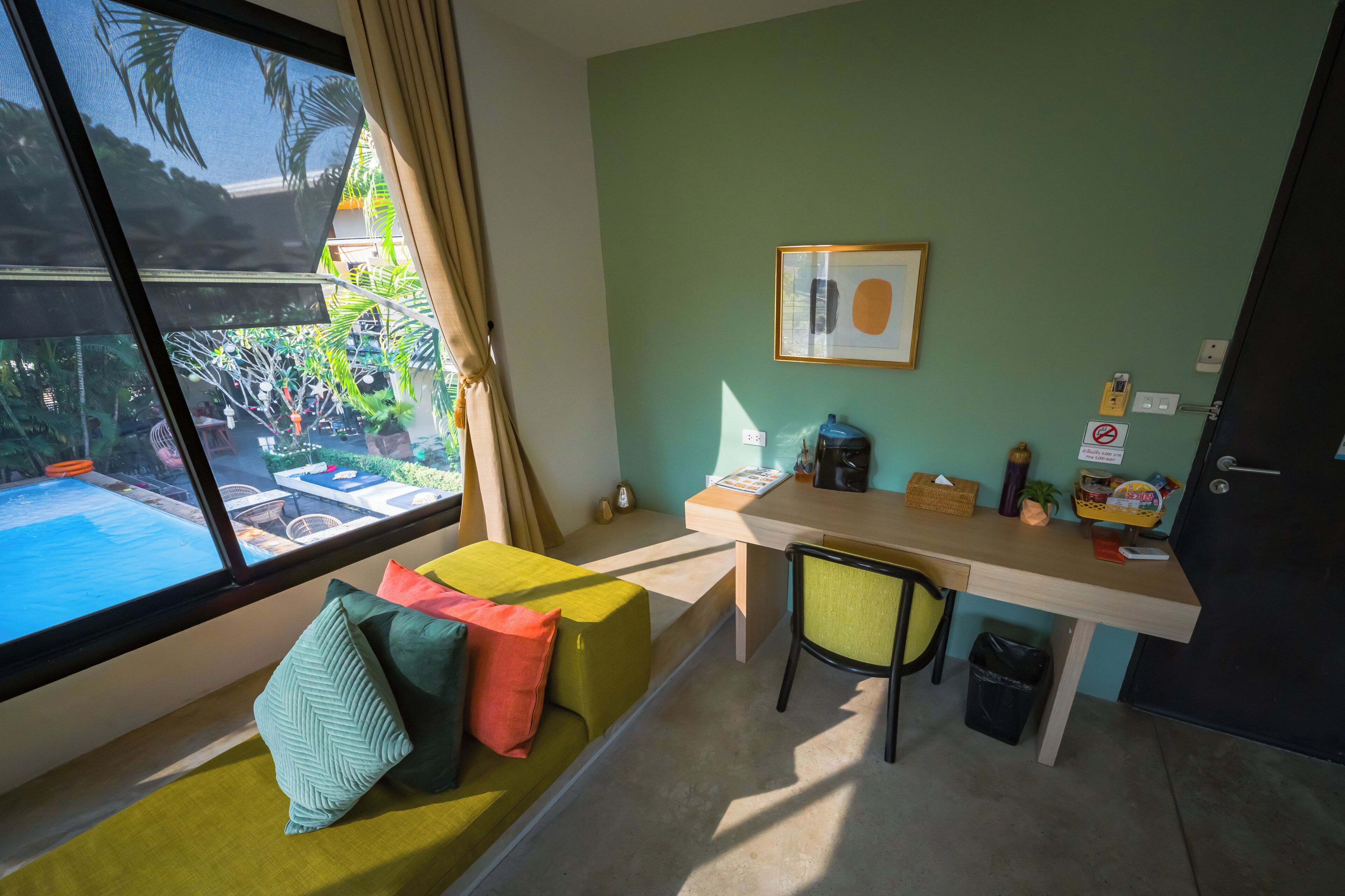 A room with a view of a swimming pool, a desk with a chair, a framed picture on the wall, and a trash bin.