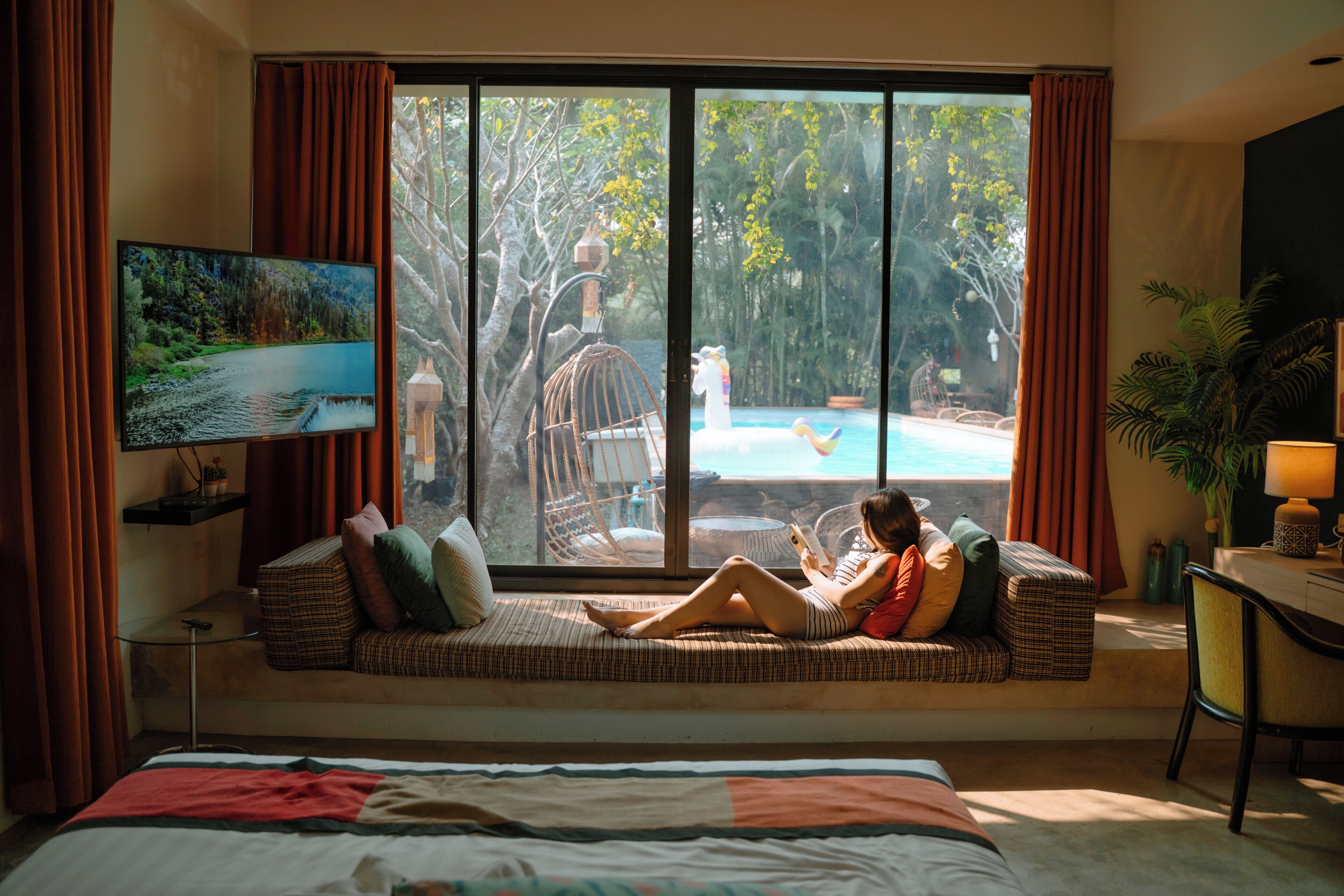 A room with a large window, a bed, a sofa with cushions, a television, and a desk with a lamp.