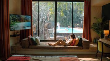 A room with a large window, a bed, a sofa with cushions, a television, and a desk with a lamp.