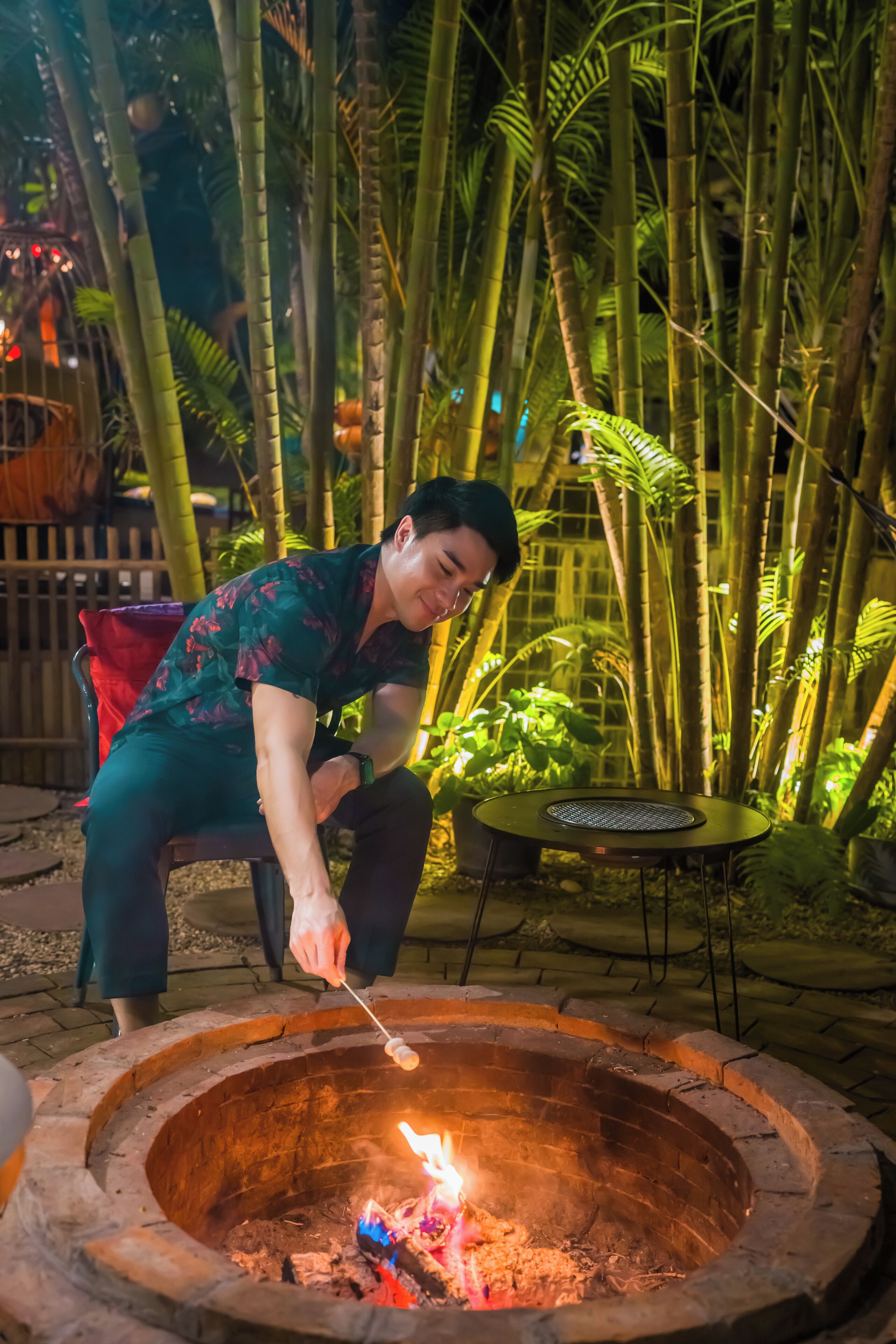 A person roasting marshmallows over a fire pit in an outdoor setting with bamboo plants.
