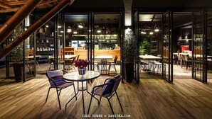 Un área de comedor al aire libre con mesas y sillas redondas, rodeada de paredes de vidrio y asientos en el interior.
