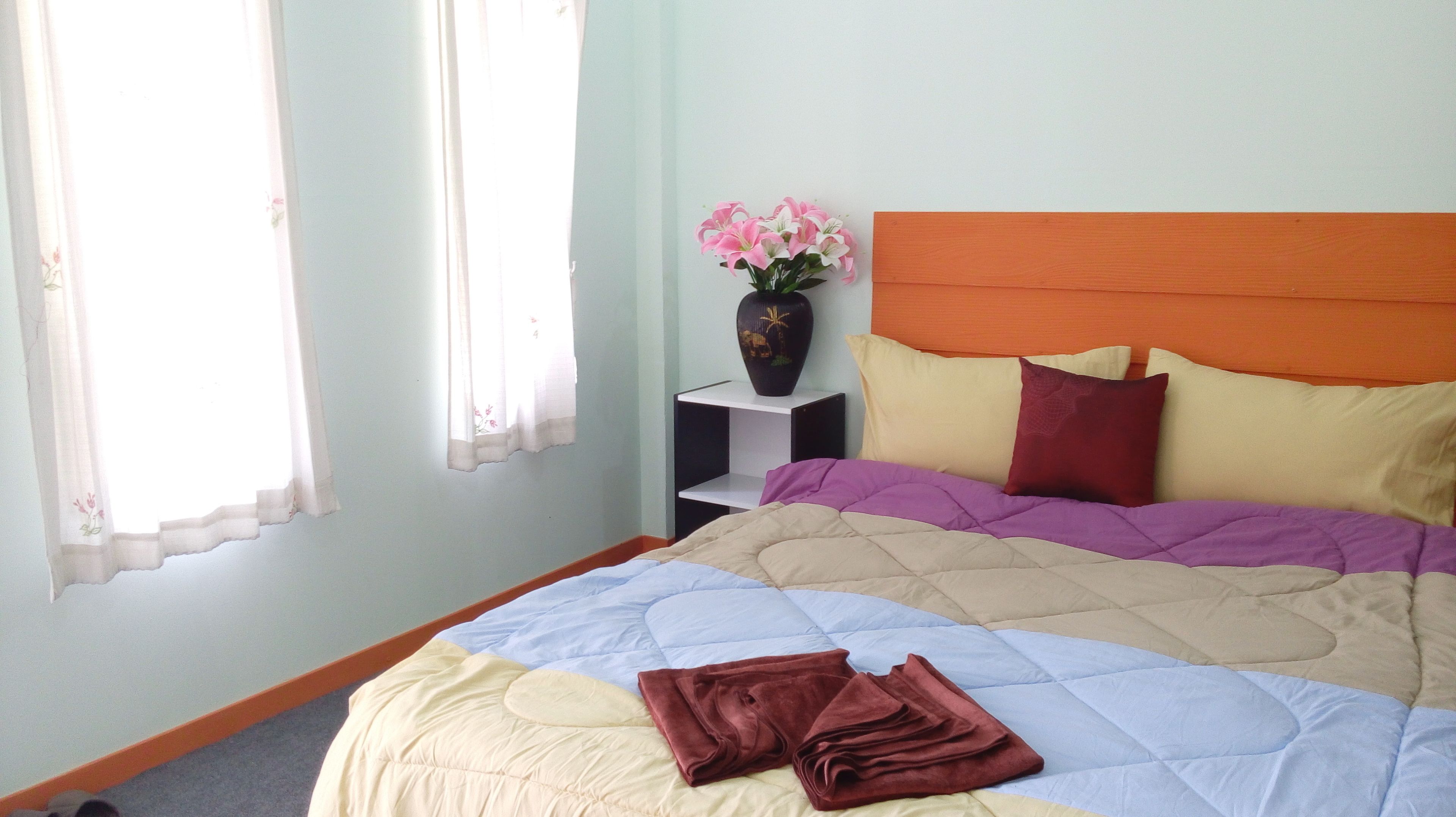 A bedroom with a bed, pillows, a nightstand, a vase with flowers, and a window with curtains.