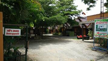 Un village rural thaïlandais avec des maisons traditionnelles en bois, un chemin de gravier et un panneau indiquant les coordonnées.