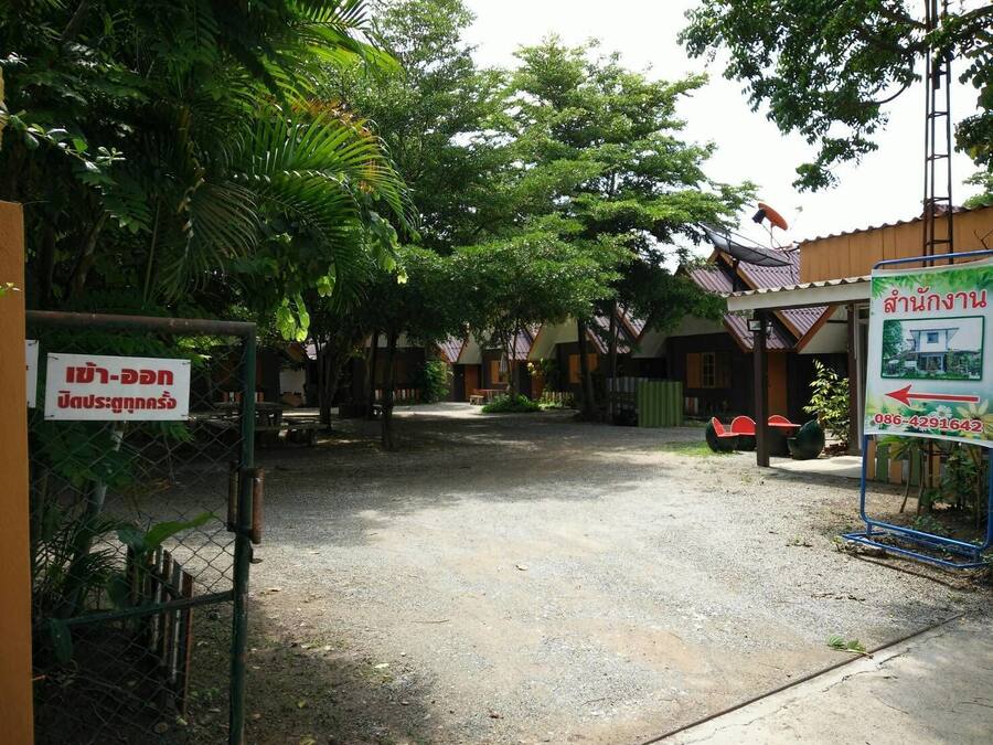 Un village rural thaïlandais avec des maisons traditionnelles en bois, un chemin de gravier et un panneau indiquant les coordonnées.