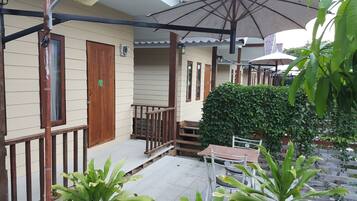 Un espacio al aire libre techado con una escalera de madera que conduce a un porche, rodeado de vegetación y una mesa con sillas.