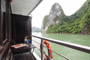 A table with a magazine, a drink, and a life preserver on a boat with a view of a green river and a large cliff.