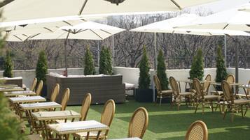 An outdoor patio with tables and chairs under large umbrellas, surrounded by greenery and a stone wall.