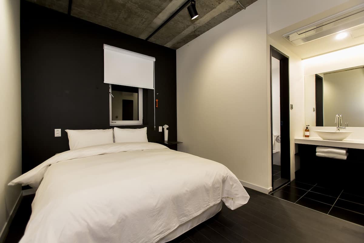 A modern hotel room with a large bed, a bedside table, a mirror, and a bathroom with a sink and mirror.