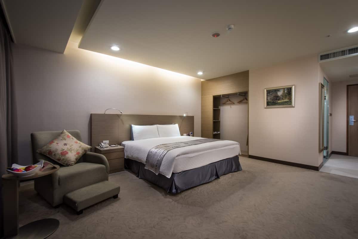 A modern hotel room with a bed, a chair, a bedside table, a wardrobe, and a painting on the wall.