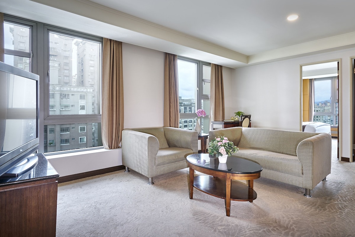 A hotel room with two sofas, a coffee table, a TV, and large windows with curtains.