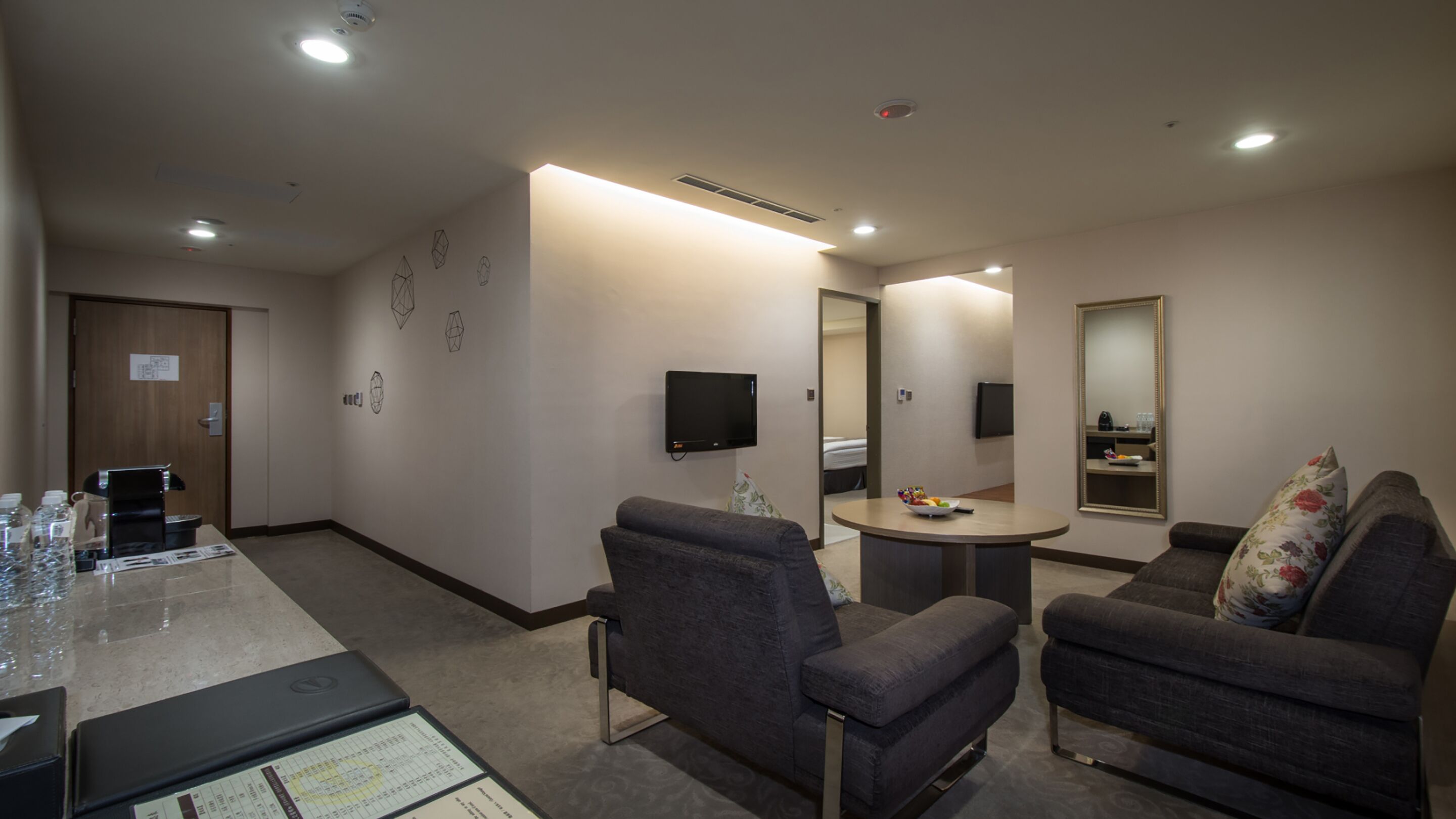A hotel room with a sofa, armchair, coffee machine, TV, and a small table with a bowl of fruit.