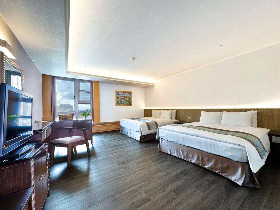 A hotel room with a large bed, a smaller bed, a desk with a TV, a chair, and a window with a city view.