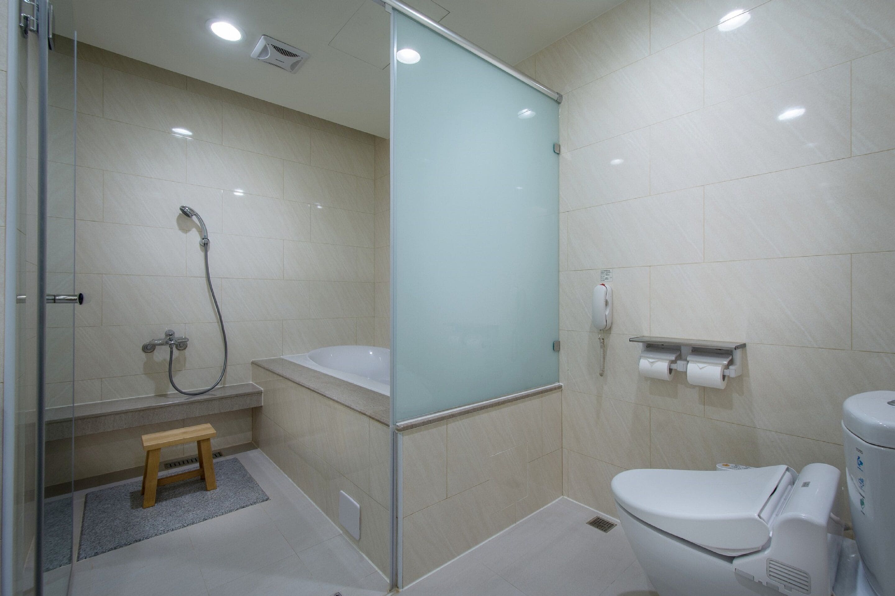 Un baño moderno con cabina de ducha de vidrio, bañera, inodoro y un pequeño taburete de madera.