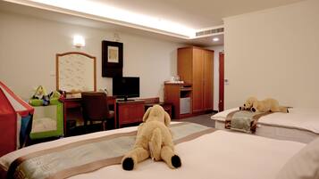 Habitación de hotel con cama, escritorio con computadora, silla, televisor y un oso de peluche sobre la cama.