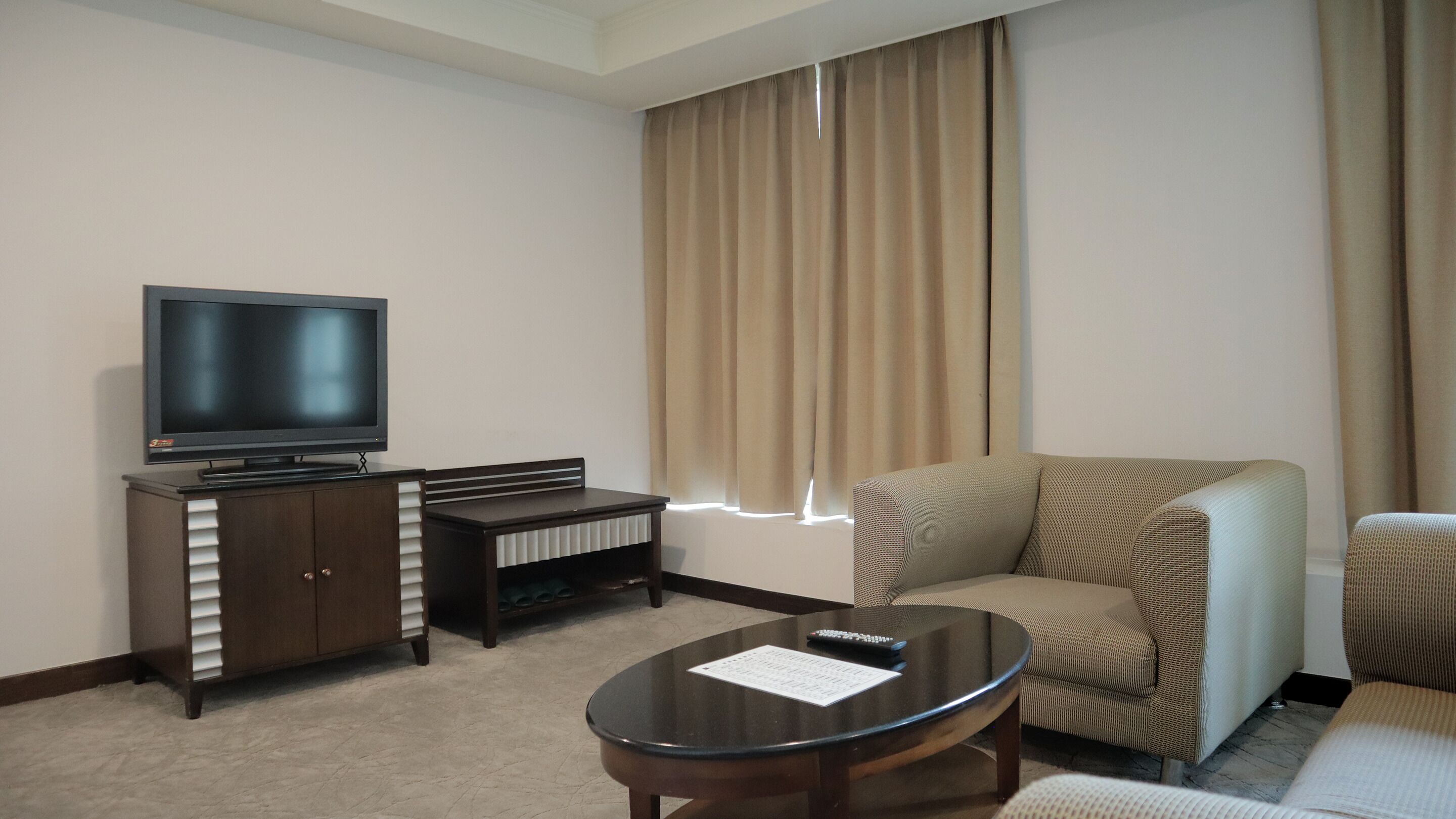 Habitación de hotel con televisión de pantalla plana, un pequeño armario de madera, una mesita de centro redonda y un sofá color beige.