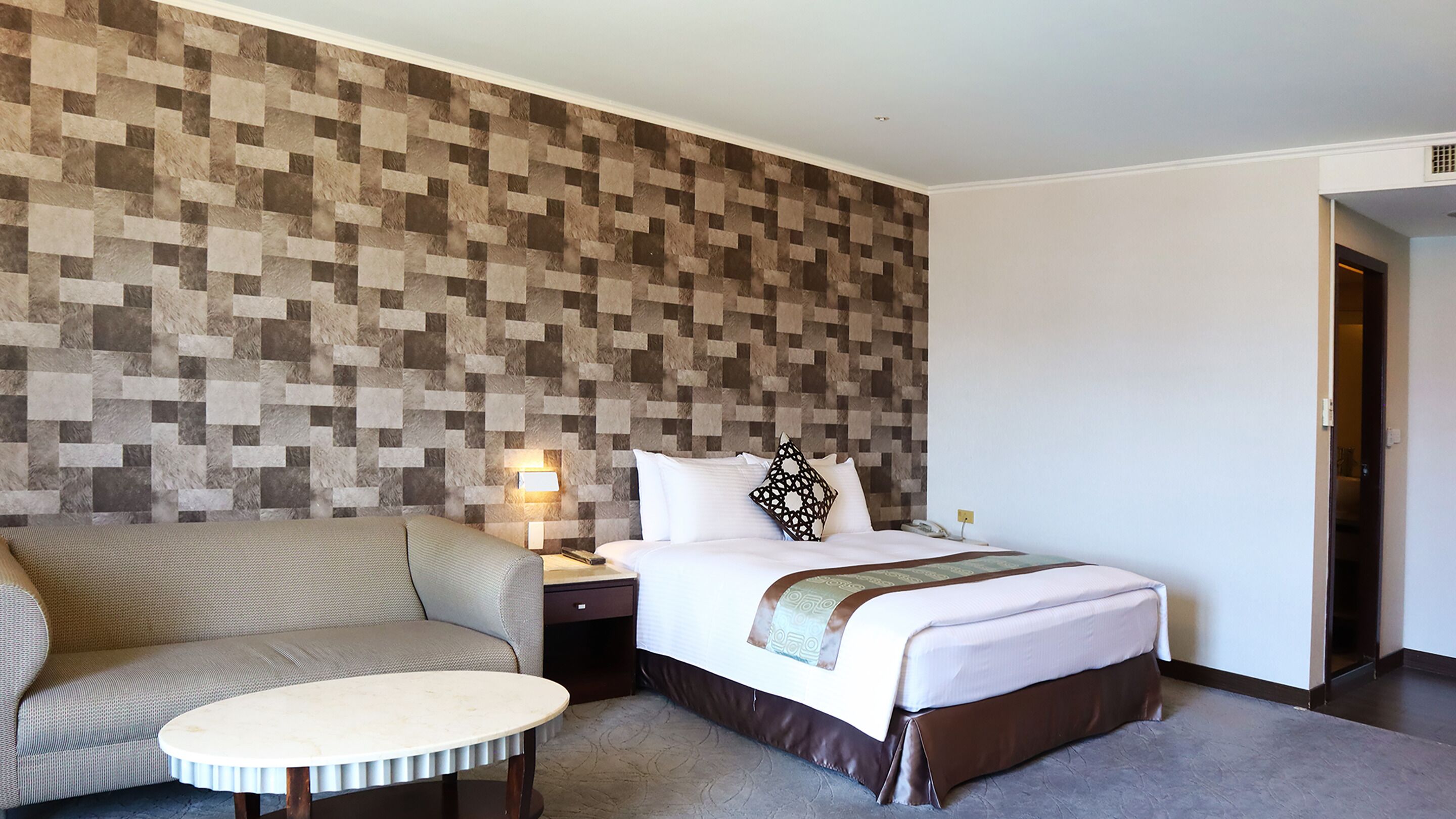 A hotel room with a bed, a sofa, a small table, a nightstand, and a wall with a geometric pattern.