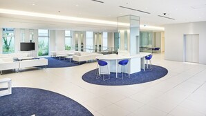 Un lobby moderno y espacioso con una zona de estar central, un televisor de pantalla plana y grandes ventanales que ofrecen vistas a zonas verdes.