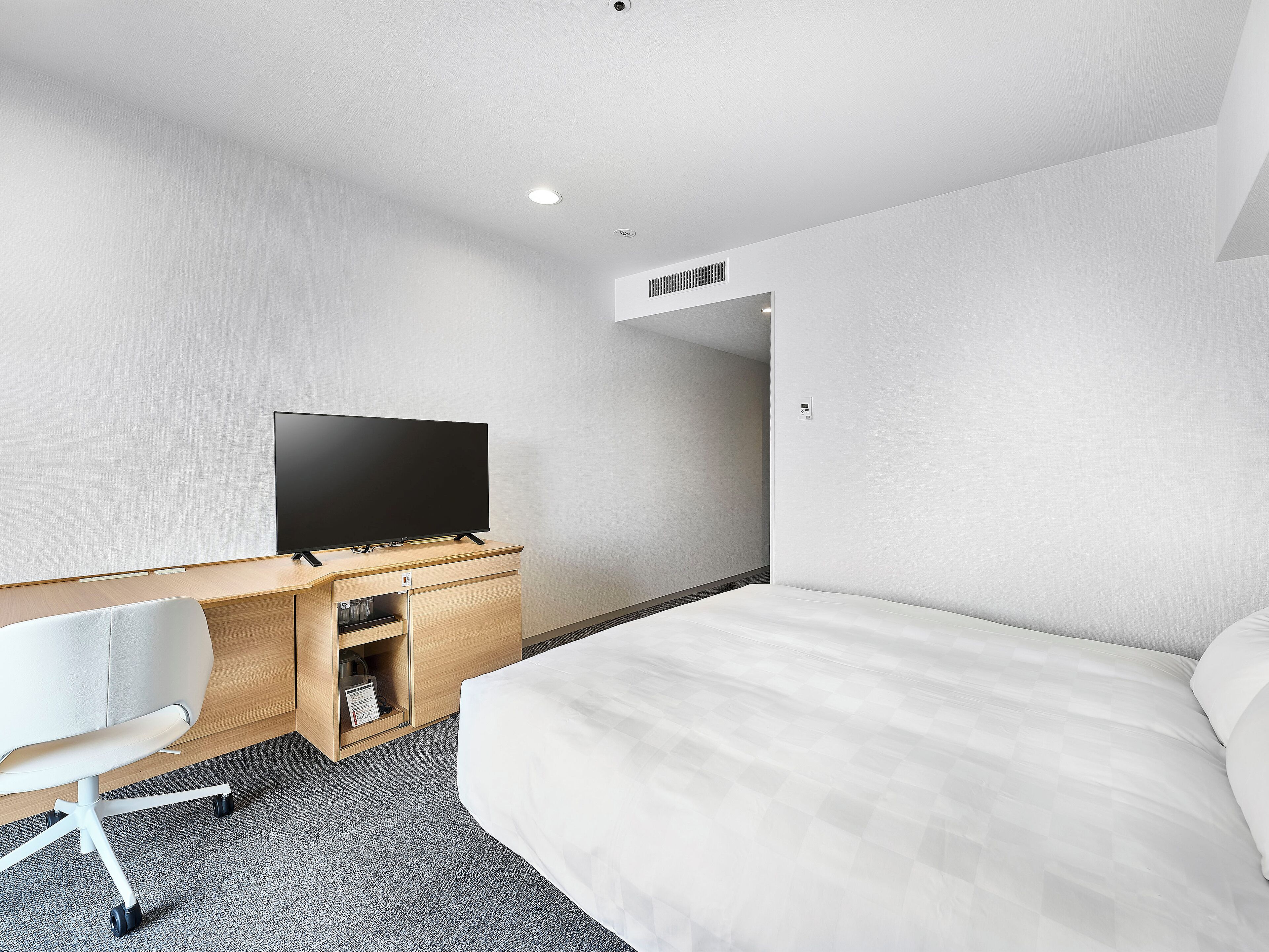 A hotel room with a bed, a desk with a flat-screen TV, a chair, and a carpeted floor.
