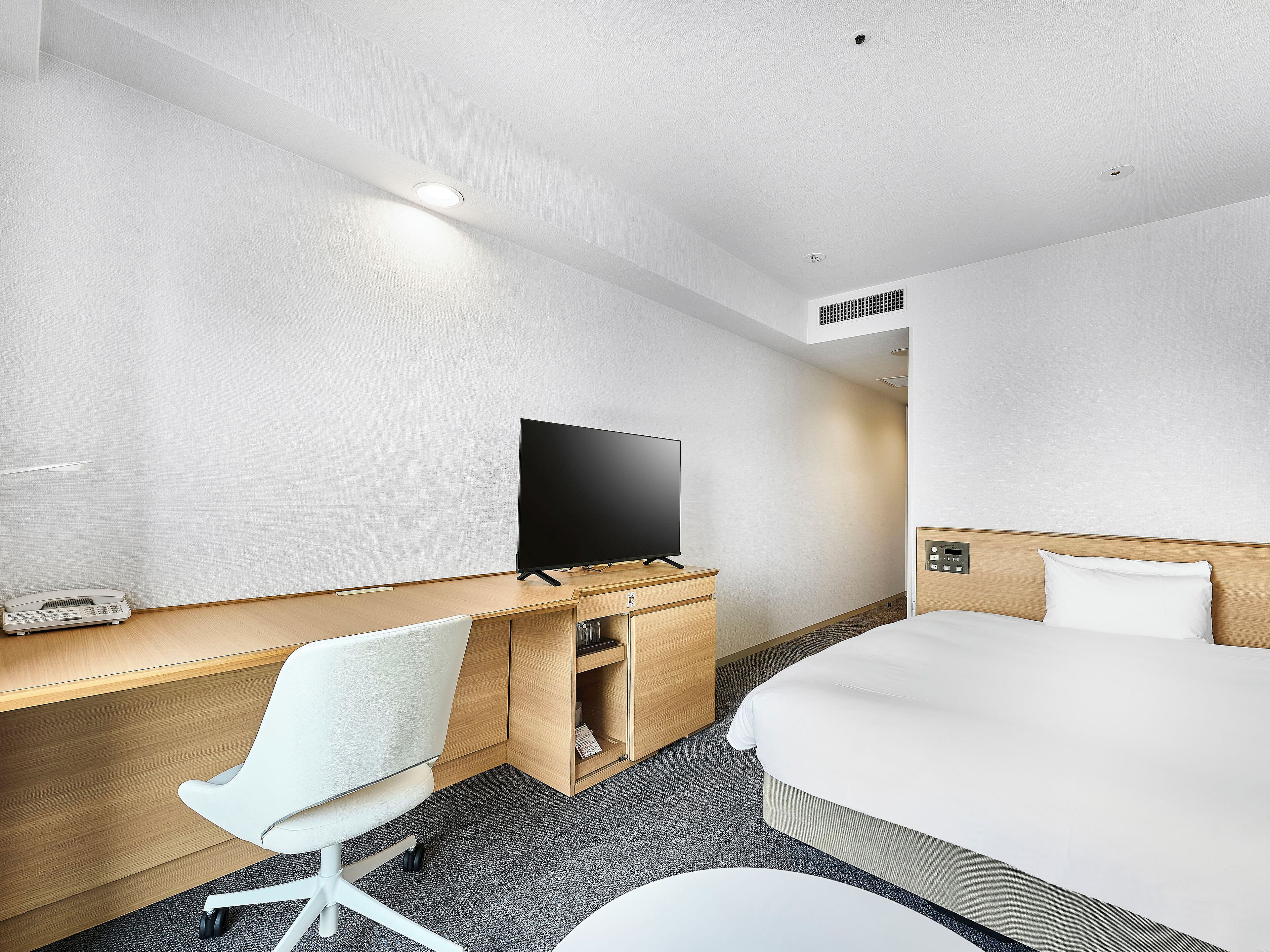A hotel room with a bed, a desk with a flat-screen TV, a chair, and a telephone.