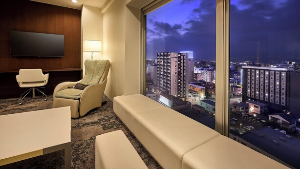 Habitación de hotel con un ventanal grande, un televisor de pantalla plana, un sofá, una silla y una pequeña mesa.