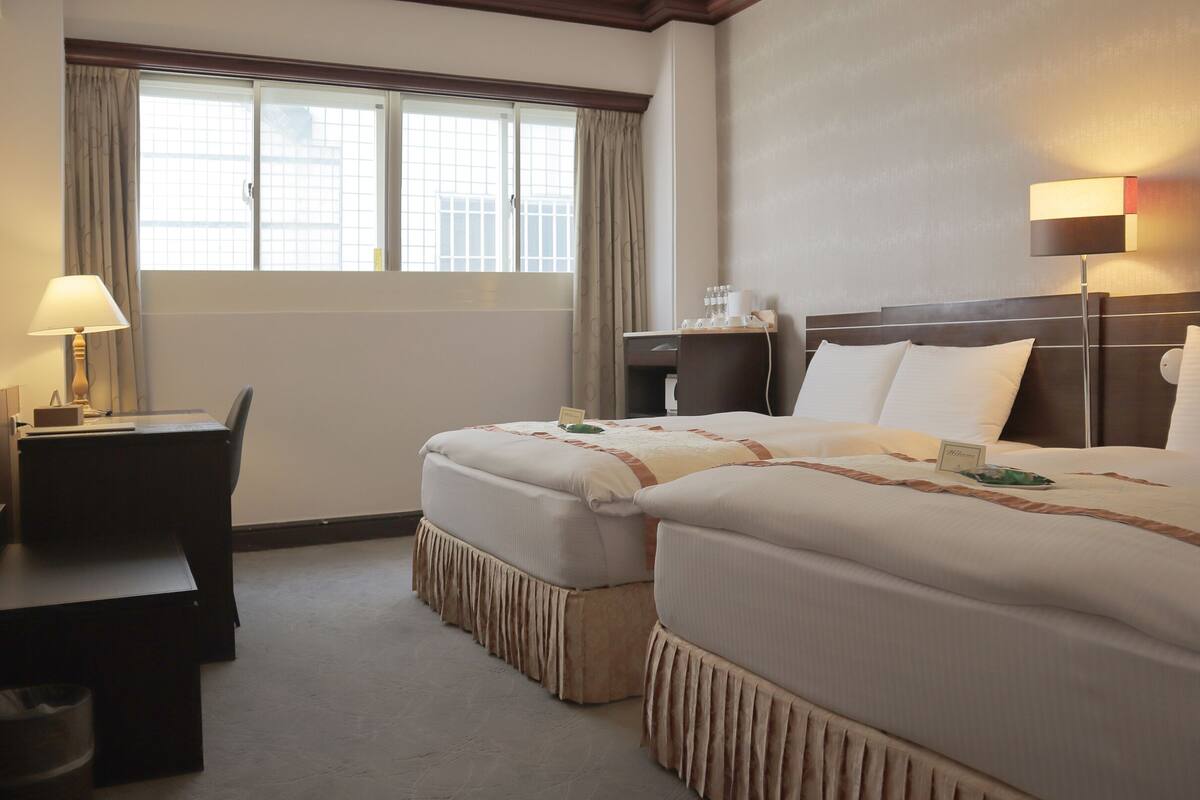 A hotel room with two beds, a desk, a chair, a nightstand, and a window with curtains.
