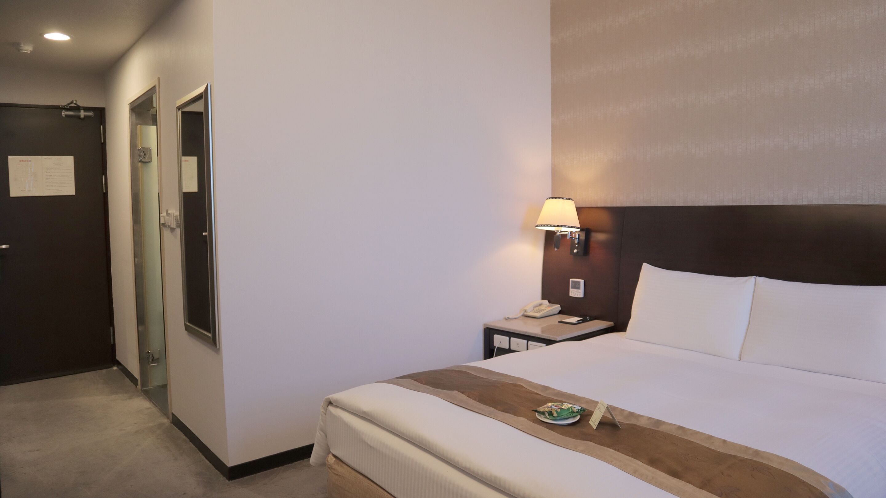 A hotel room with a bed, bedside table, lamp, and a door with a mirror.