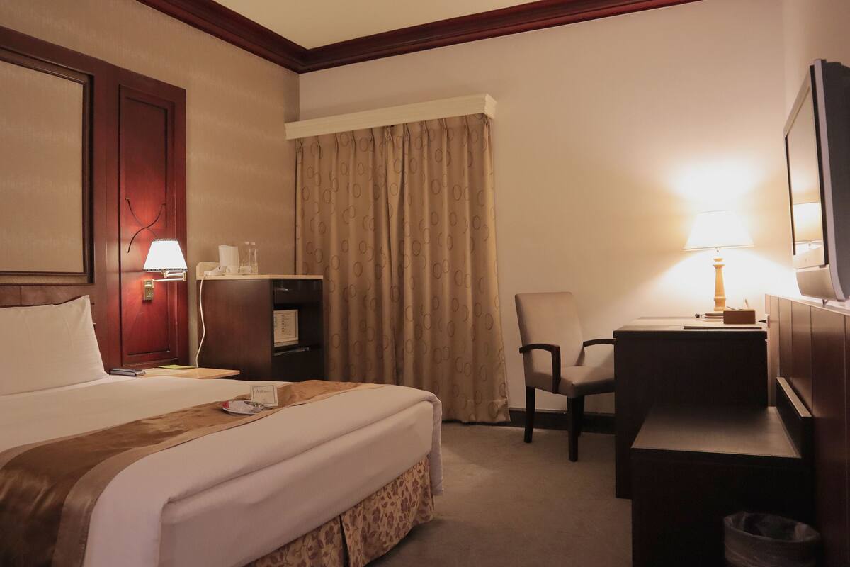 A hotel room with a large bed, a desk, a chair, a TV, and a lamp.