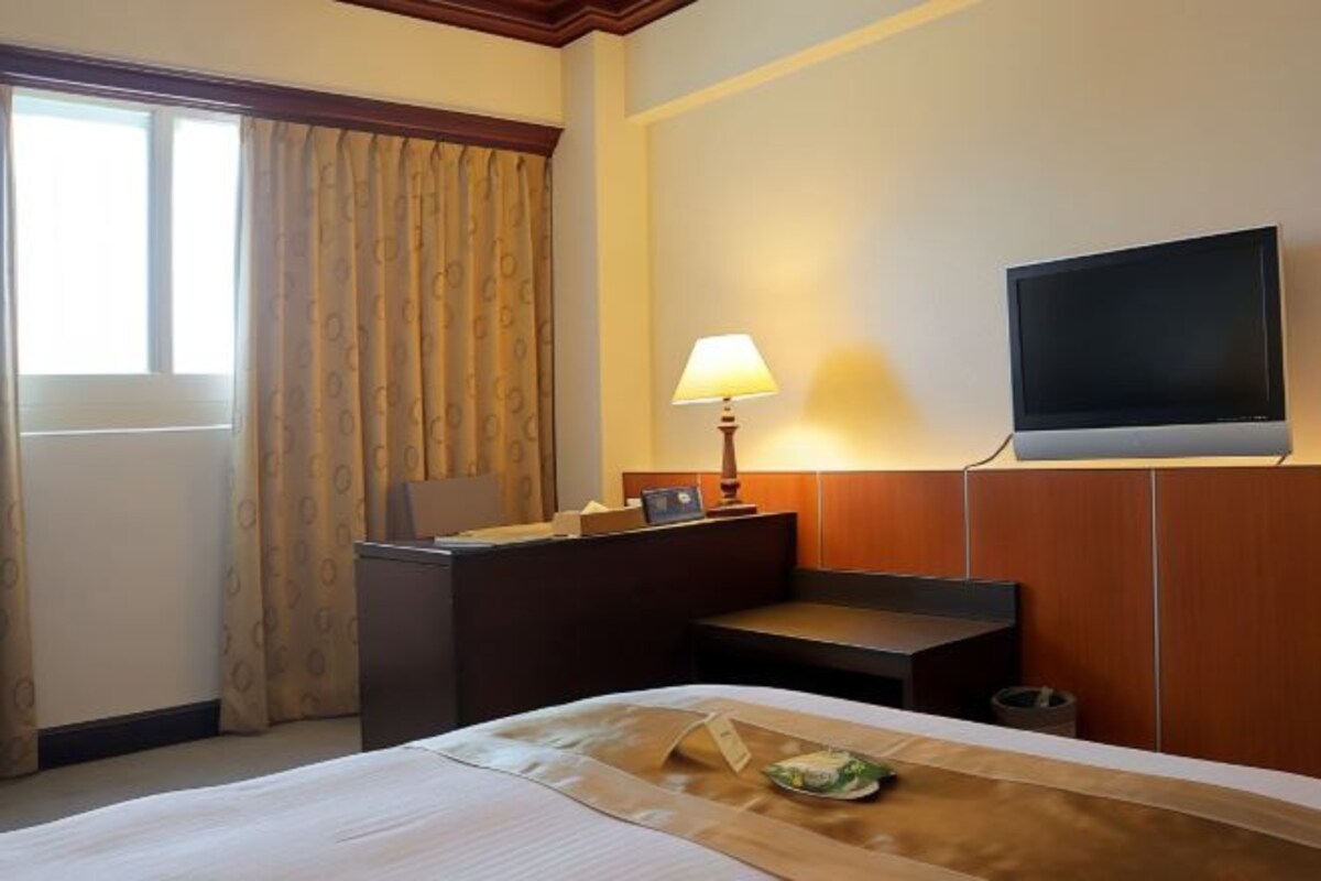 A hotel room with a bed, a desk with a lamp, a television, and a window with curtains.