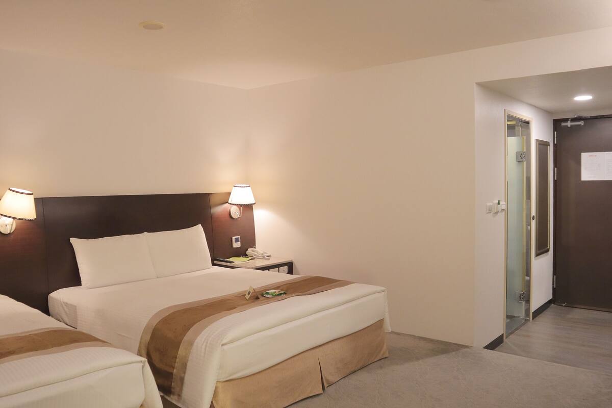 A hotel room with two beds, each with a nightstand, a lamp, and a phone. There is a door leading to another room.