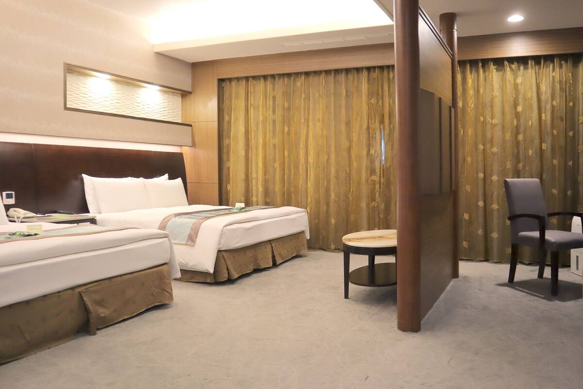 A hotel room with two beds, a desk, a chair, and a large window with curtains.
