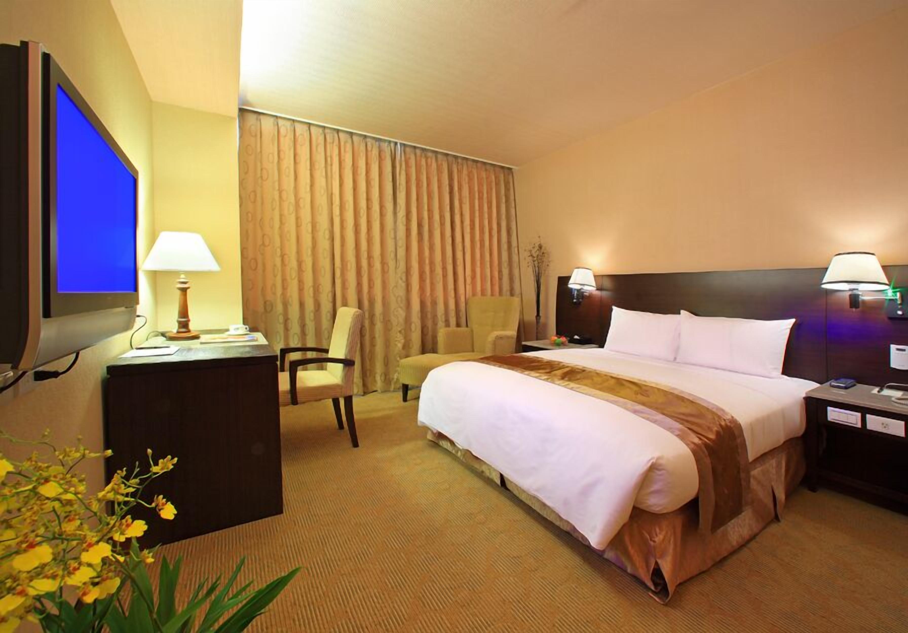 A hotel room with a bed, a desk with a lamp, a chair, a television, and a nightstand with a lamp.