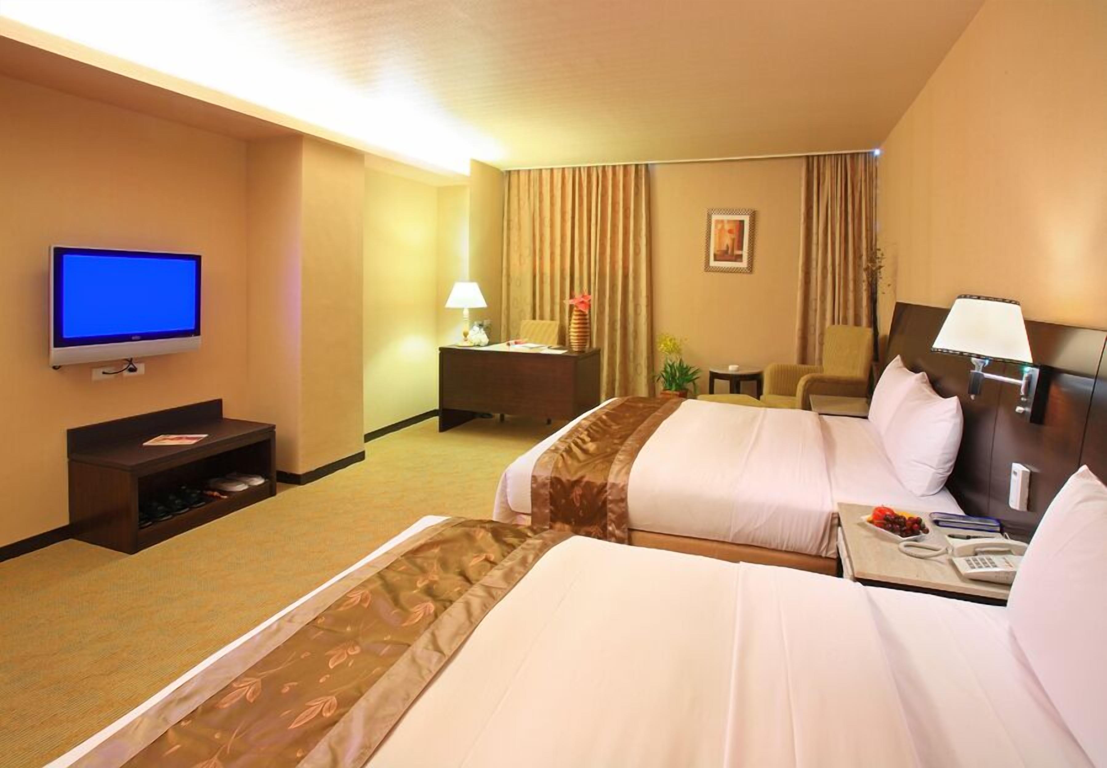 A hotel room with two beds, a television, a desk with a lamp, and a small table with a phone and a fruit basket.