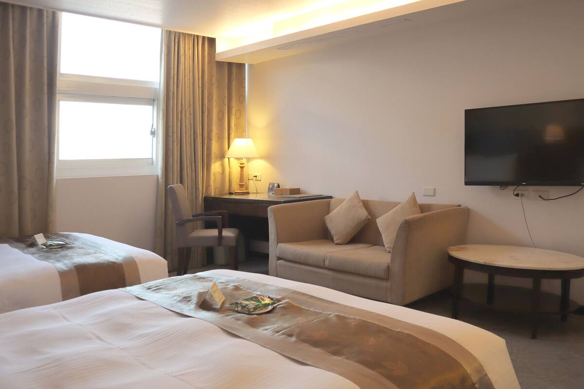 A hotel room with two beds, a desk with a lamp, a TV, and a small table.