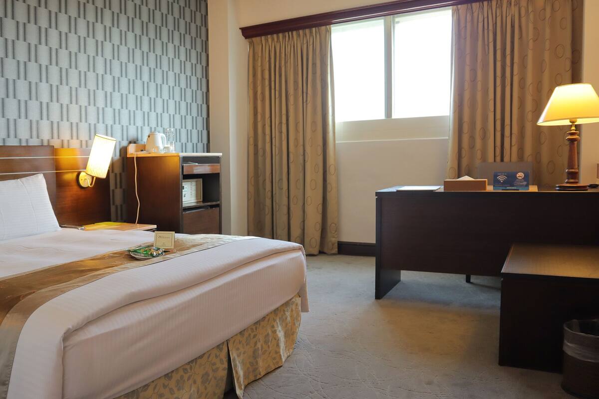 A hotel room with a neatly made bed, a desk with a lamp, a nightstand with a phone and a lamp, a window with curtains, and a wall with a geometric pattern.