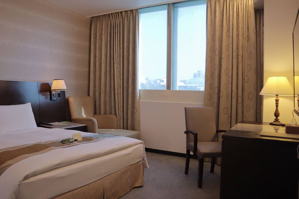 A hotel room with a large bed, a desk, a chair, and a window with curtains.