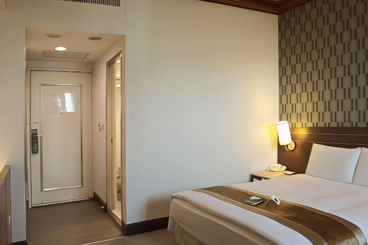 A hotel room with a bed, bedside table, lamp, and a door leading to another room.