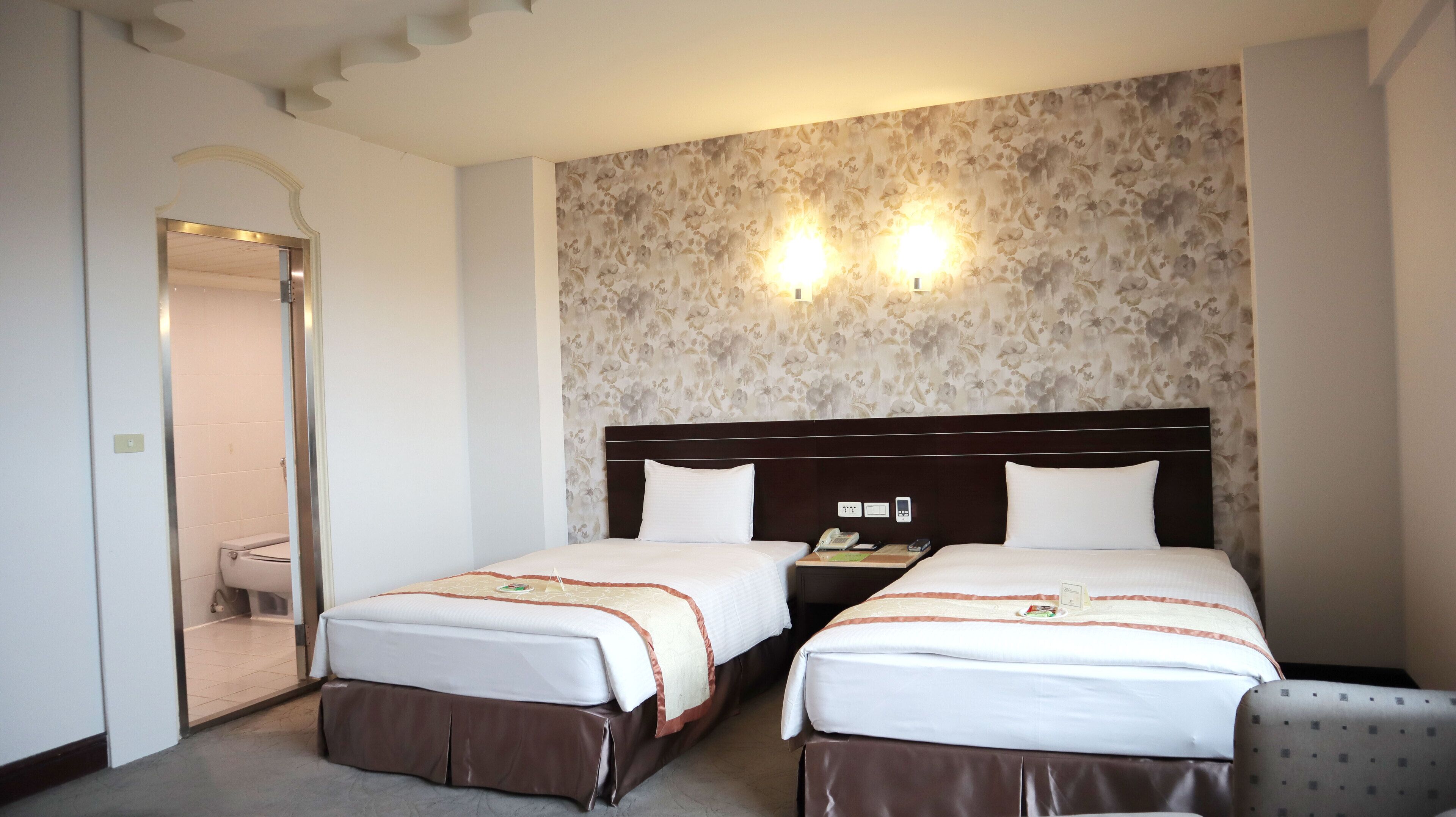 A hotel room with two beds, a wooden headboard, a floral wallpaper, and a small bedside table with a lamp.