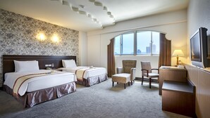 Et hotelværelse med to senge, et skrivebord, en stol, et fjernsyn og et stort vindue med udsigt over byen.