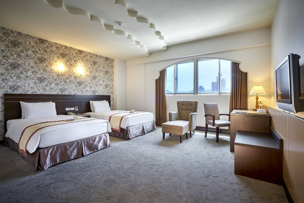 A hotel room with two beds, a desk, a chair, a television, and a large window with a city view.