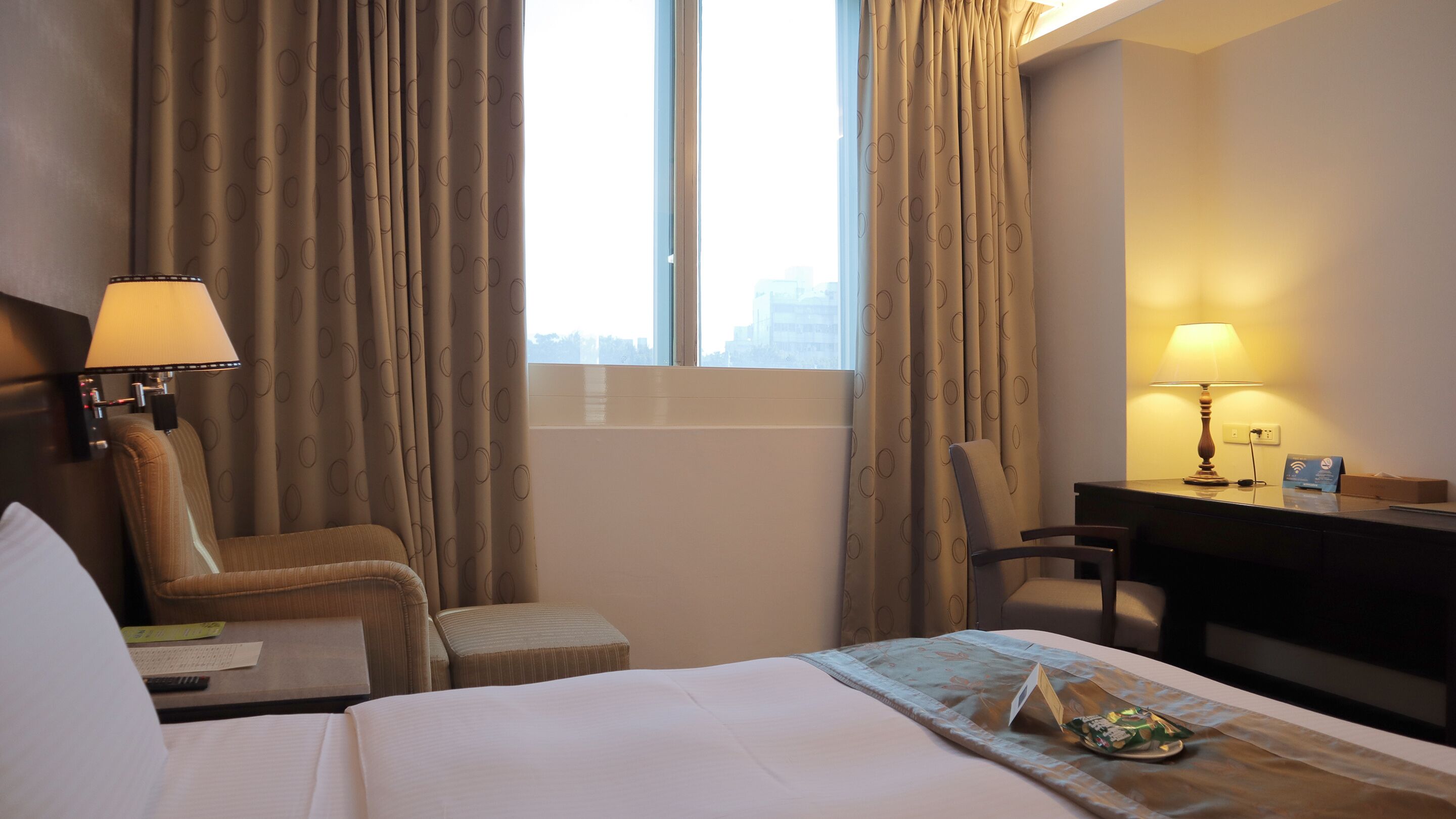 Een hotelkamer met een bed, twee stoelen, een bureau, een lamp en een raam met gordijnen.