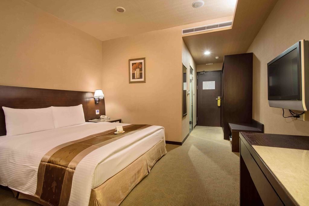A hotel room with a large bed, a bedside table with a lamp, a television, and a door leading to a hallway.
