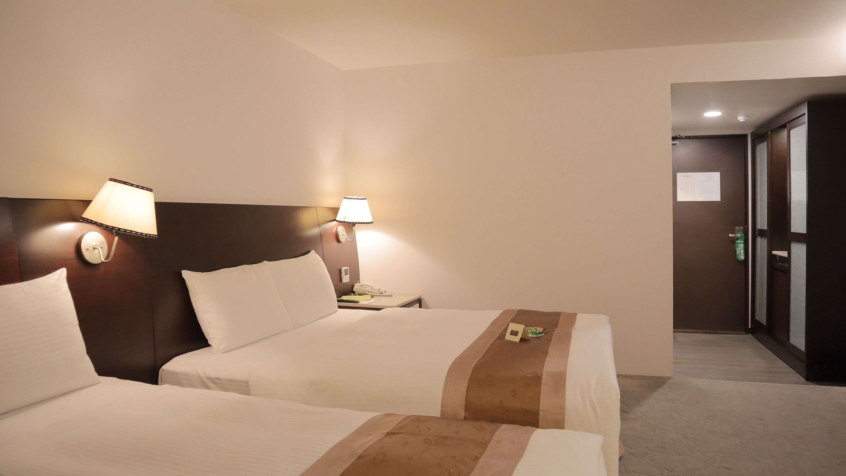 A hotel room with two beds, a wooden headboard, bedside lamps, a nightstand, and a door with a green handle.