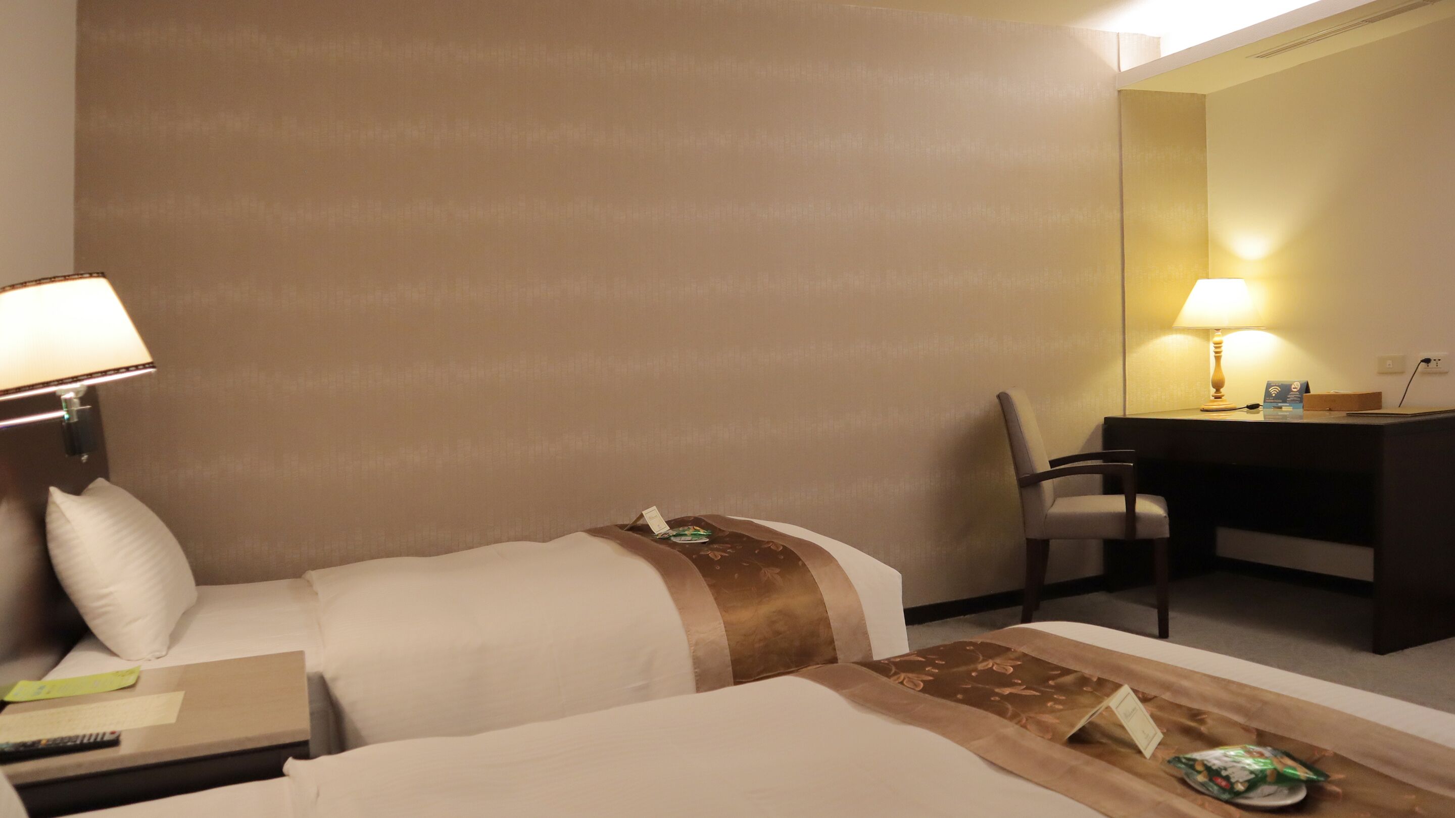 A hotel room with two beds, a desk, a chair, and a lamp.
