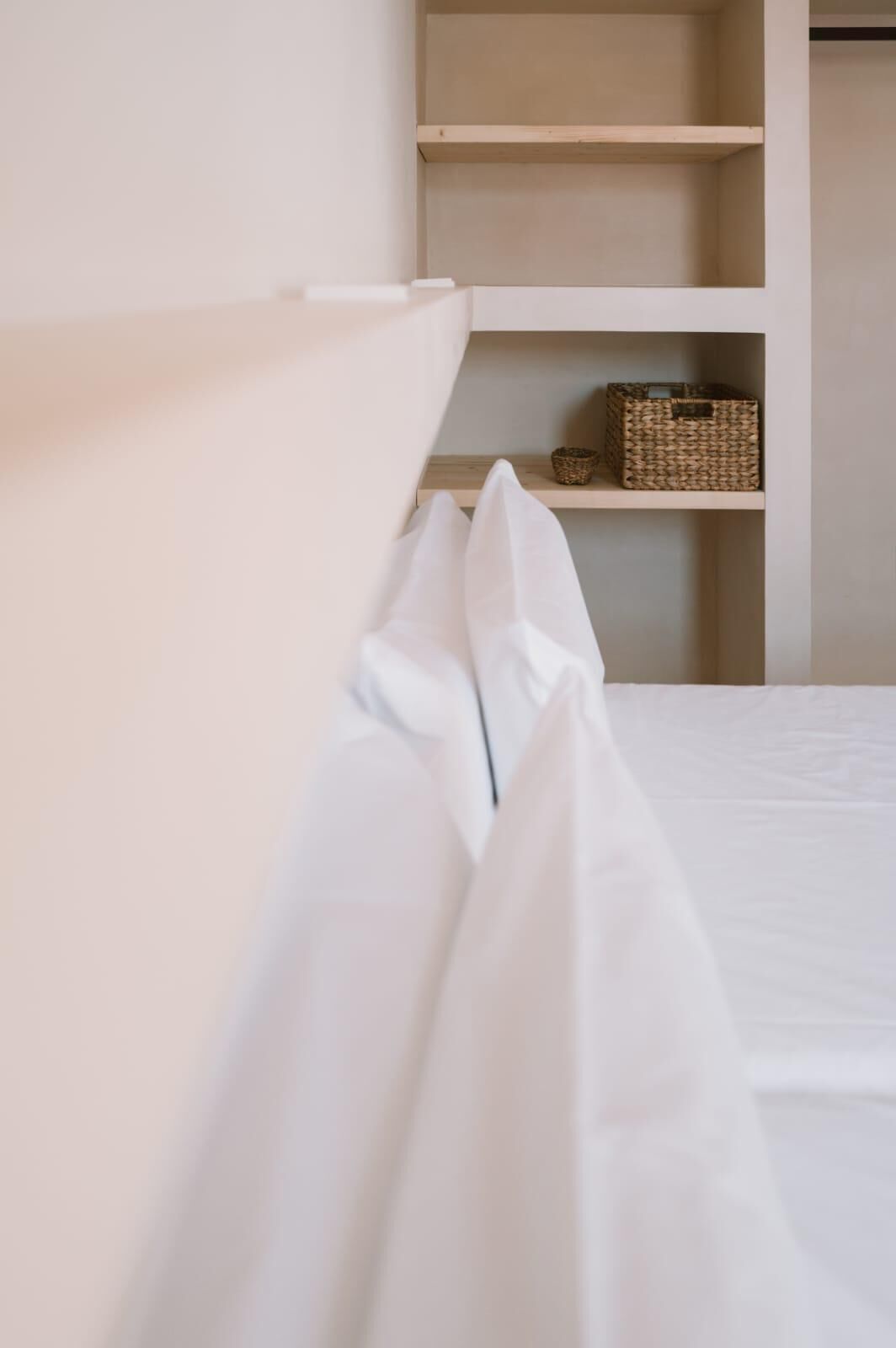 A bed with white bedding and a headboard, a shelf with baskets, and a wall with a light color.