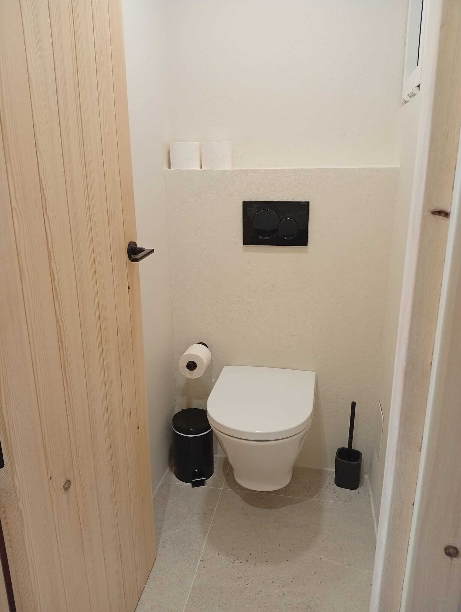 A small bathroom with a toilet, a trash bin, a toilet paper holder, and a black electrical outlet.