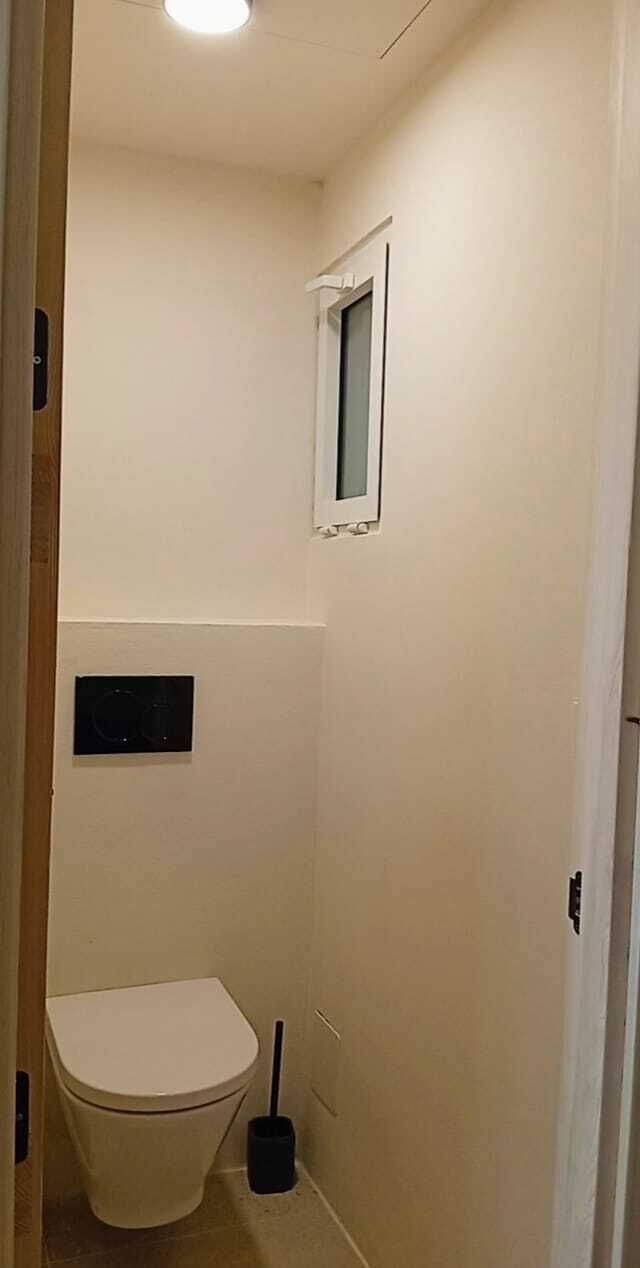 A small, narrow bathroom with a toilet, a small window, and a wall-mounted flush handle.