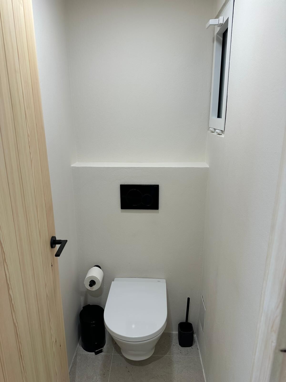 Casa de banho pequena e branca com sanita, rolo de papel higiénico, um caixote do lixo preto e uma escova de limpeza preta.
