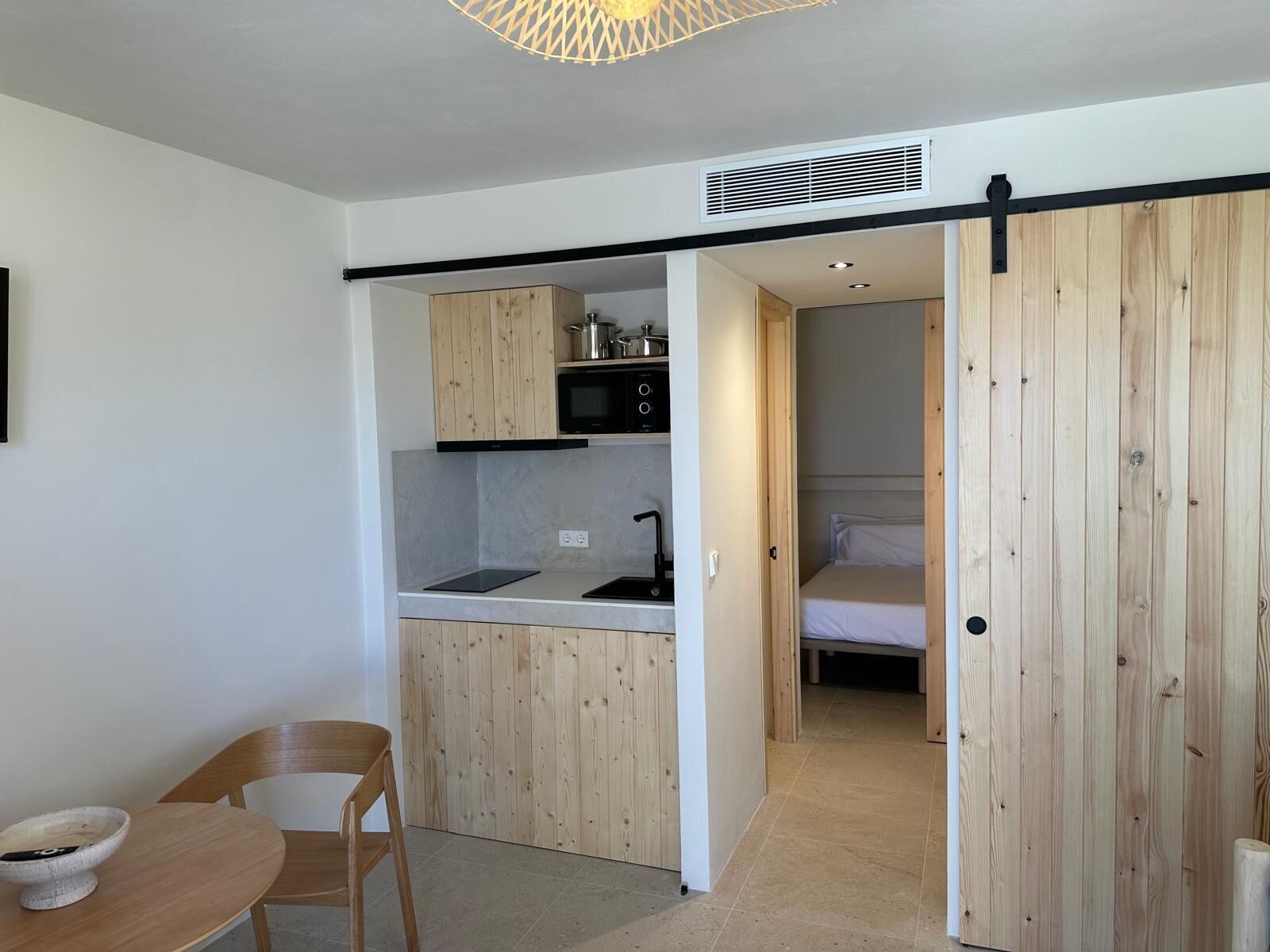A compact kitchen with wooden cabinets, a microwave, and a sink. There's a dining area with a table and chair, and a bed in the adjacent room.